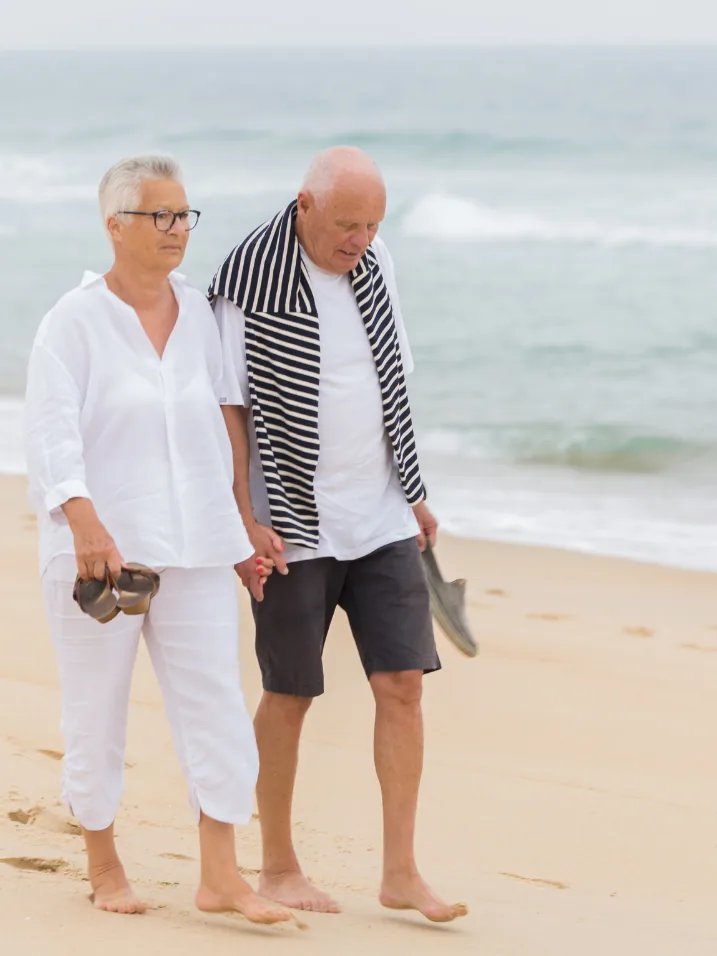 An older couple walking barefoot on the beach, holding hands